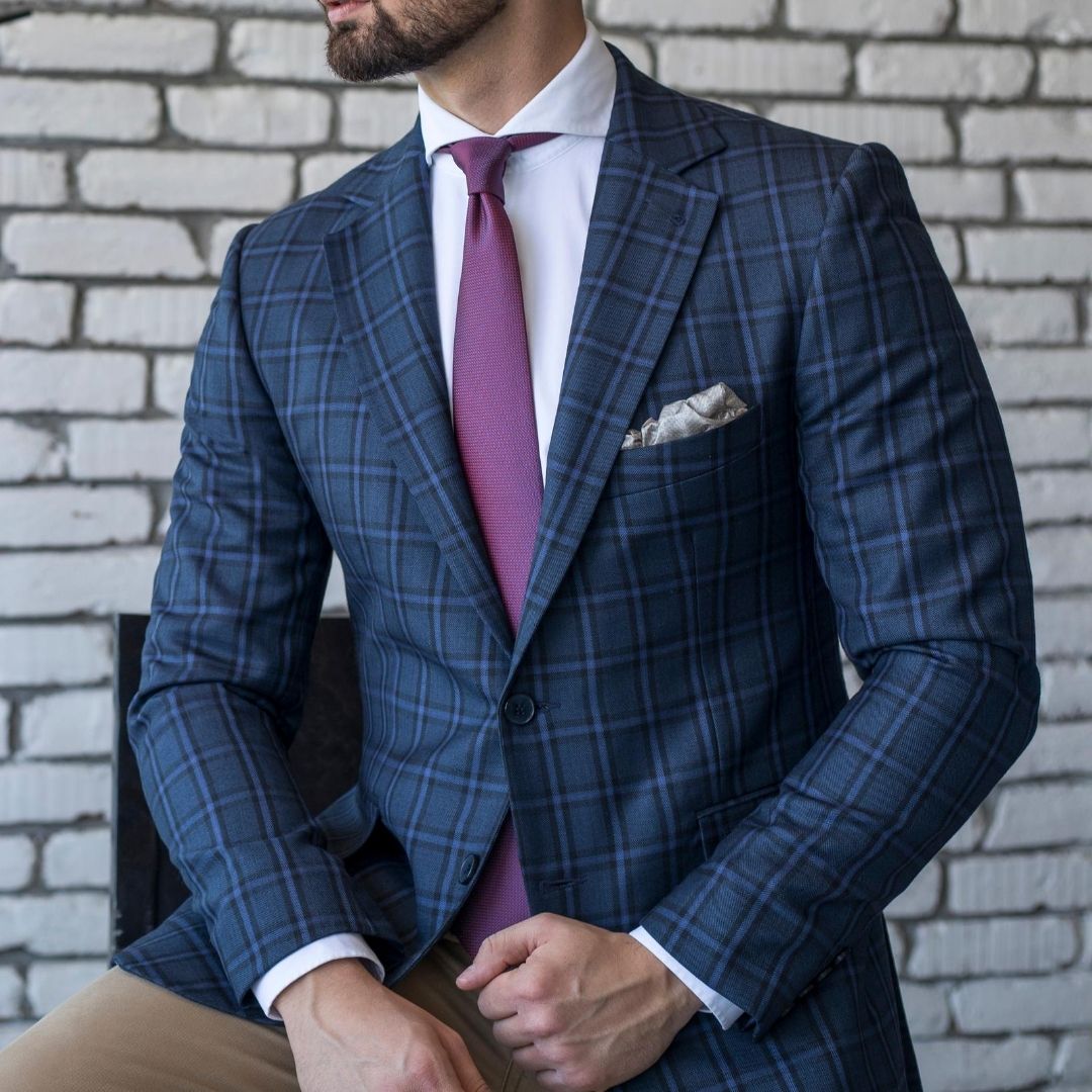 A person in a blue plaid suit with a pink tie and pocket square, sitting against a white brick wall.
