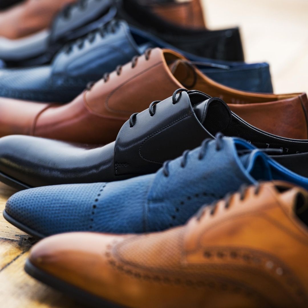 A row of men’s dress shoes in various colors and styles, including brown, black, and blue, displayed on a wooden surface.