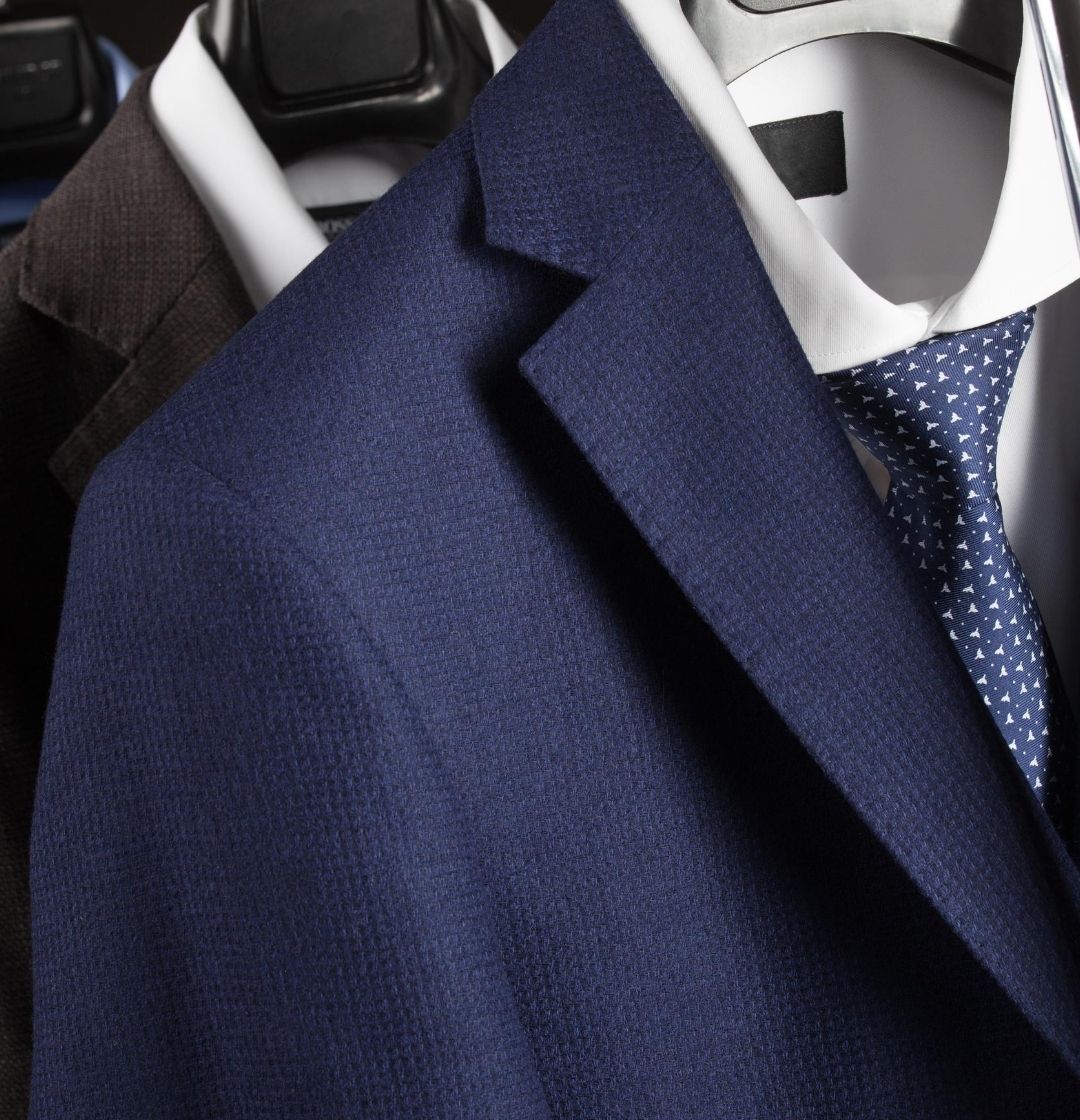 Two suits hang on hangers. The front suit is blue with a matching tie, while the back suit is dark grey.