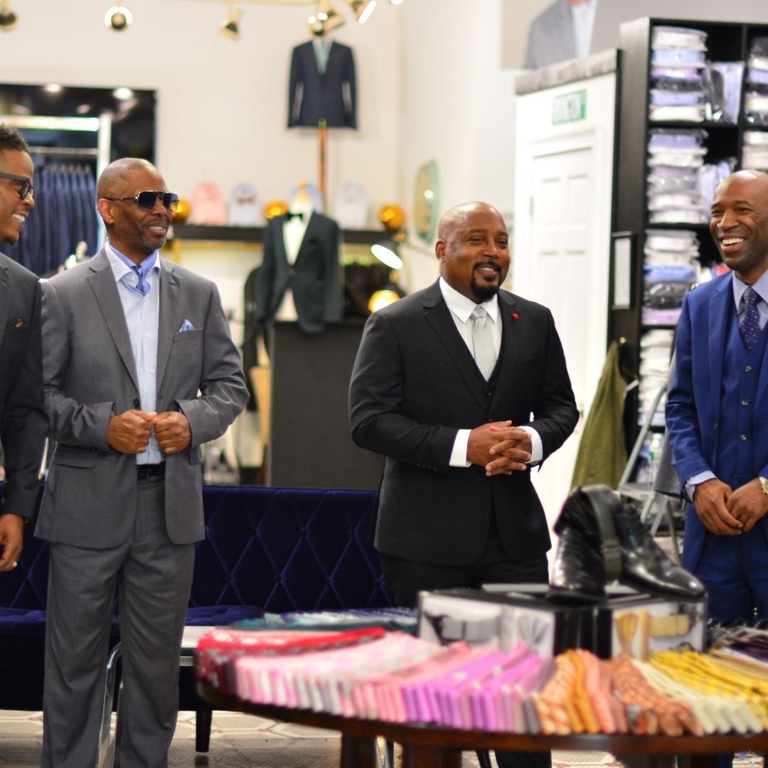 Four men in suits stand together in a clothing store, smiling and engaging in conversation. Shelves with neatly folded shirts are visible in the background.