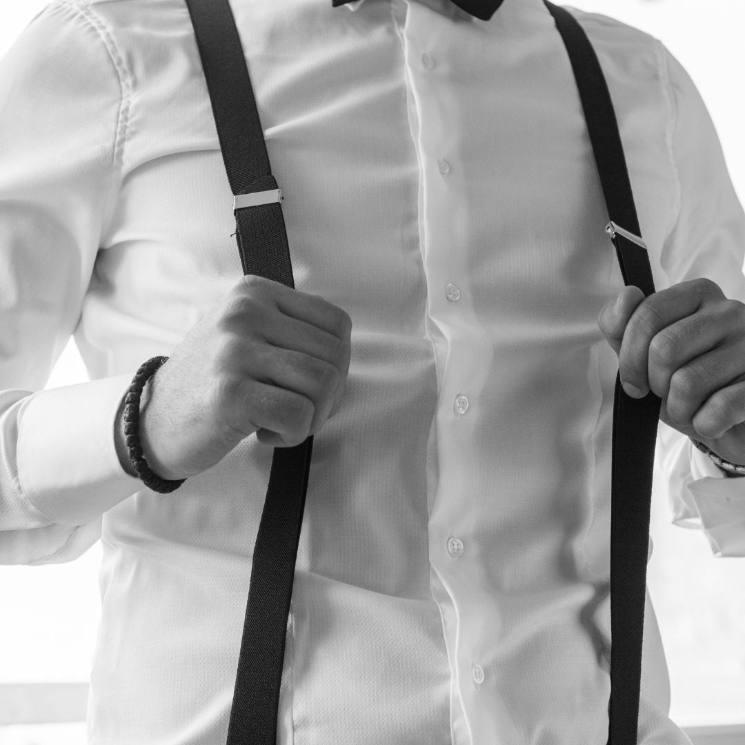 A man adjusts his black suspenders over a white dress shirt, focusing on the suspenders with a blurred background.