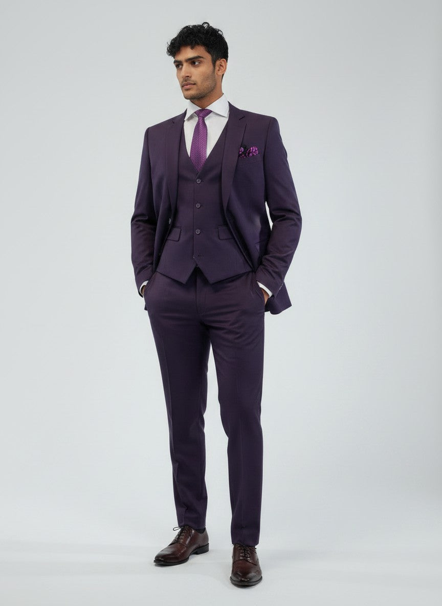 A man dressed in the FUBU Purple Vested Suit by Fubu The Colle, paired with a white shirt, purple tie, and dress shoes, stands with hands in pockets against a plain light gray background.