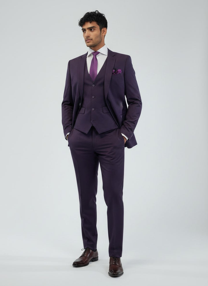 A man dressed in the FUBU Purple Vested Suit by Fubu The Colle, paired with a white shirt, purple tie, and dress shoes, stands with hands in pockets against a plain light gray background.