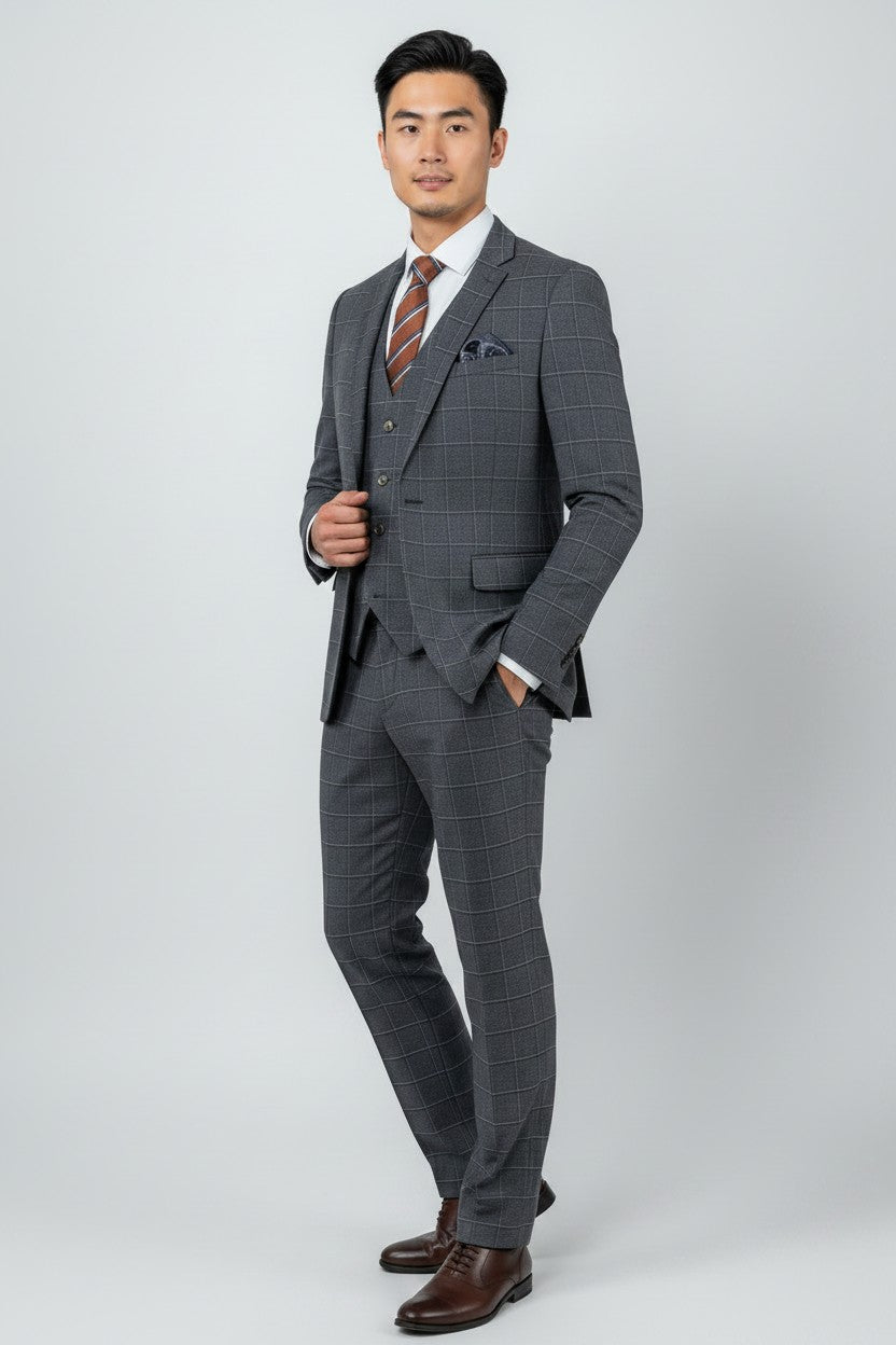 A man poses against a plain light grey background wearing the FUBU Navy Pin Check Windowpane Vested Suit by Fubu The Colle. The three-piece slim fit suit is styled with a white shirt, brown striped tie, pocket square, and brown dress shoes. For added comfort and mobility, the suit features stretch armholes.
