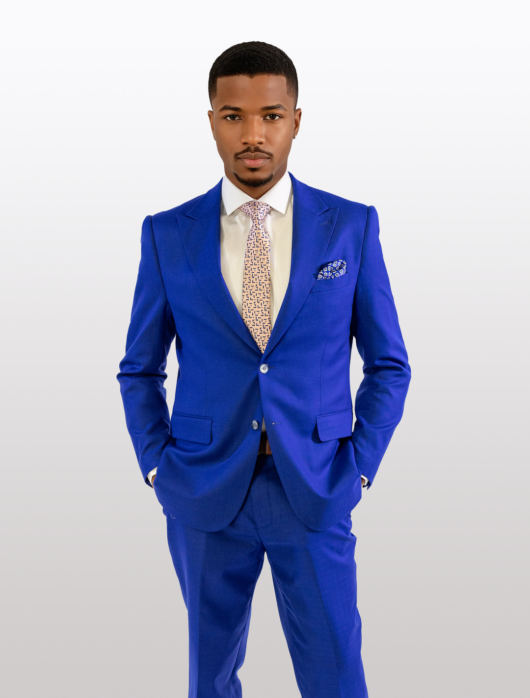A person in a FUBU Royal Blue Solid Peak Lapel 2-Piece Suit by Fubu The Colle, paired with a white shirt and patterned tie, stands with hands in pockets against a plain background.