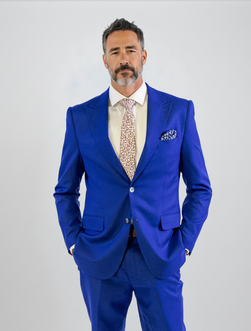 A man with short dark hair and a beard wears the FUBU Royal Blue Solid Peak Lapel 2-Piece Suit by Fubu The Colle, styled with a beige dress shirt and patterned tie, standing with hands in his pockets against a plain light background.