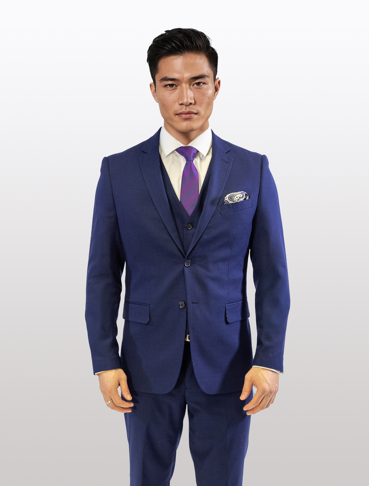 A man wearing a FUBU Berry Blue Vested Suit by Fubu The Colle, paired with a white shirt and purple striped tie, stands against a plain background.
