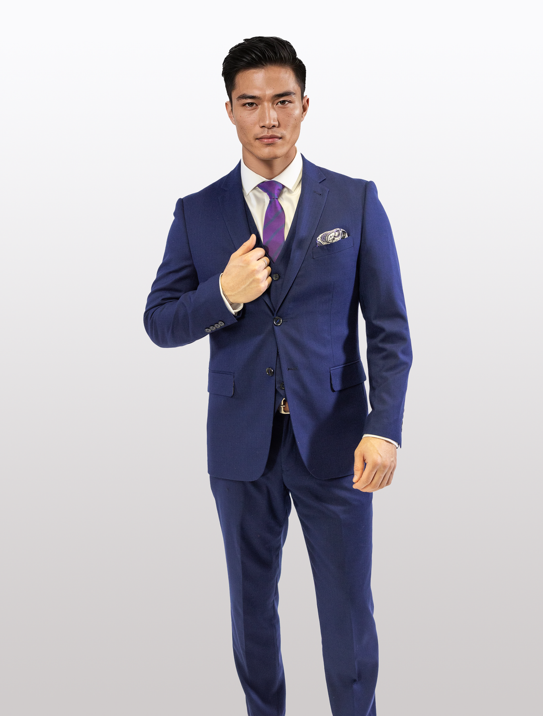 Wearing a FUBU Berry Blue Vested Suit by Fubu The Colle, an individual poses against a plain backdrop, adjusting their jacket for the perfect look with a purple tie.