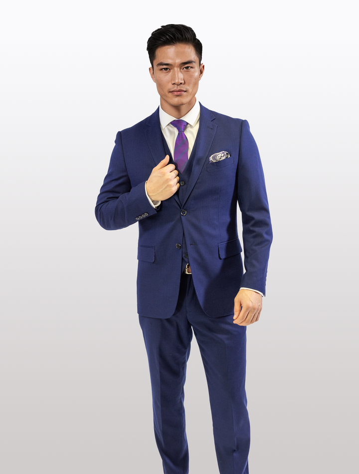 Wearing a FUBU Berry Blue Vested Suit by Fubu The Colle, an individual poses against a plain backdrop, adjusting their jacket for the perfect look with a purple tie.