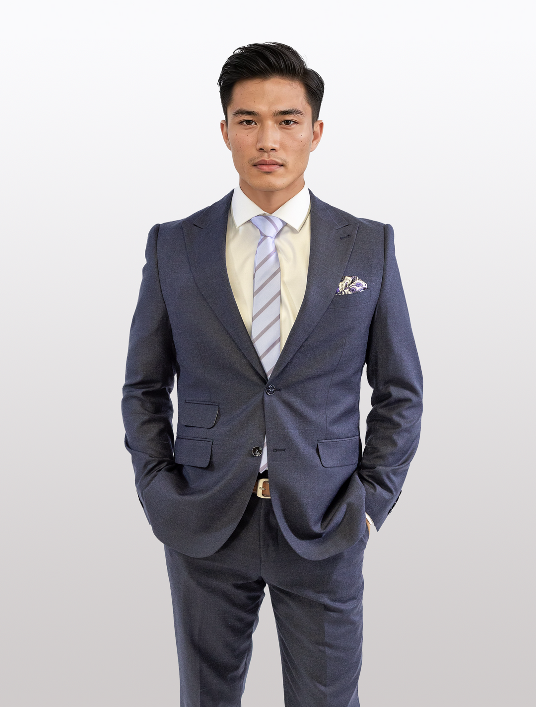 A man wearing a FUBU Polo Blue Peak Lapel 2-Piece Suit by Fubu The Colle, paired with a light yellow shirt and striped tie, stands with hands in pockets against a plain backdrop. The slim-fit suit includes a peak lapel jacket and stretch armholes for added comfort.