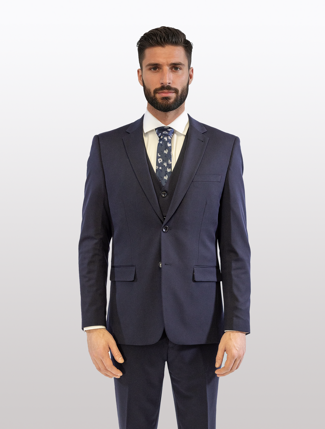 A man stands against a plain backdrop wearing the FUBU Midnight Navy with Debossed Windowpane Vested Suit by Fubu The Colle, paired with a crisp white shirt and patterned tie.