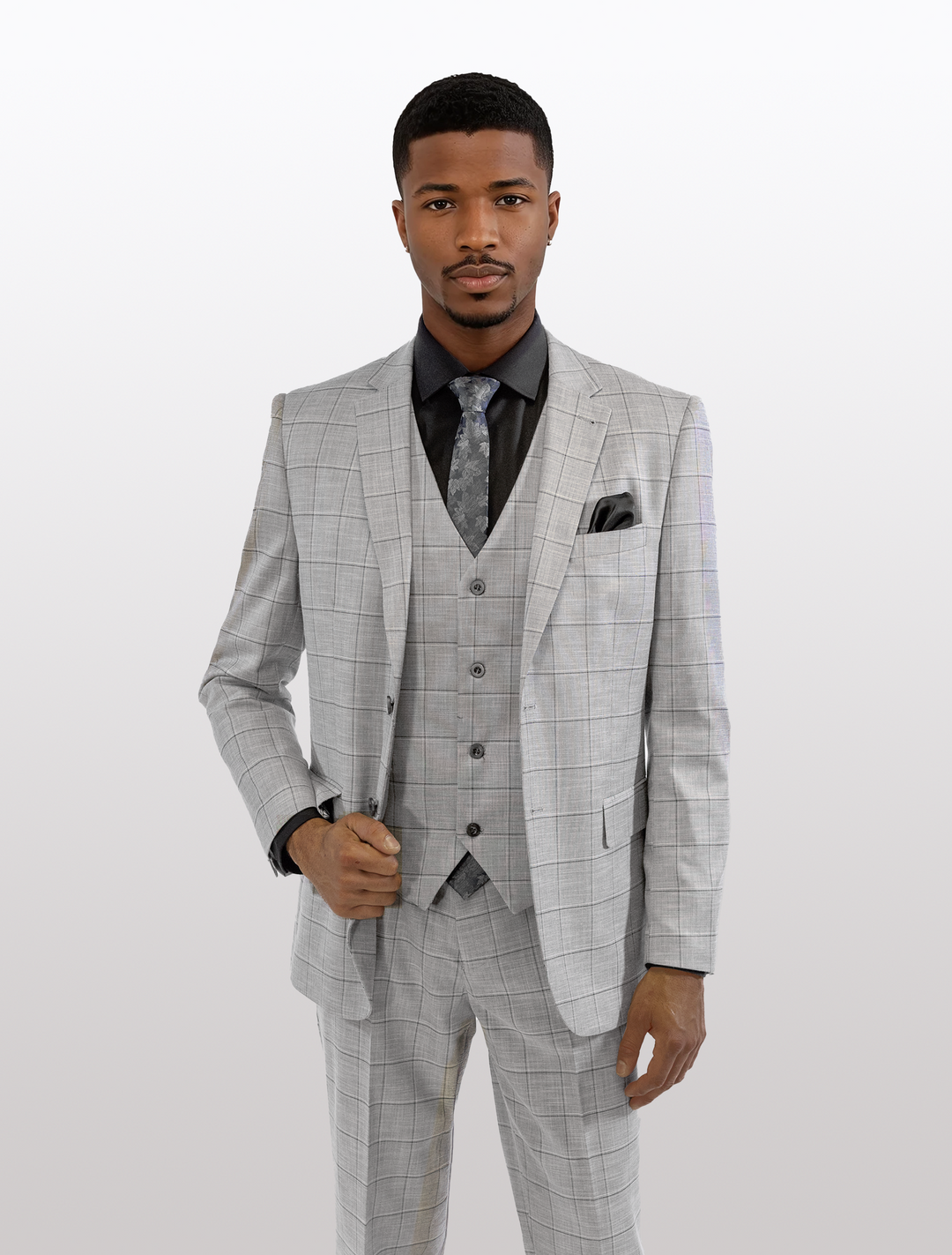 A man stands confidently in a FUBU Light Grey with Charcoal Windowpane Vested Suit by Fubu The Colle, paired with a black shirt and patterned tie.