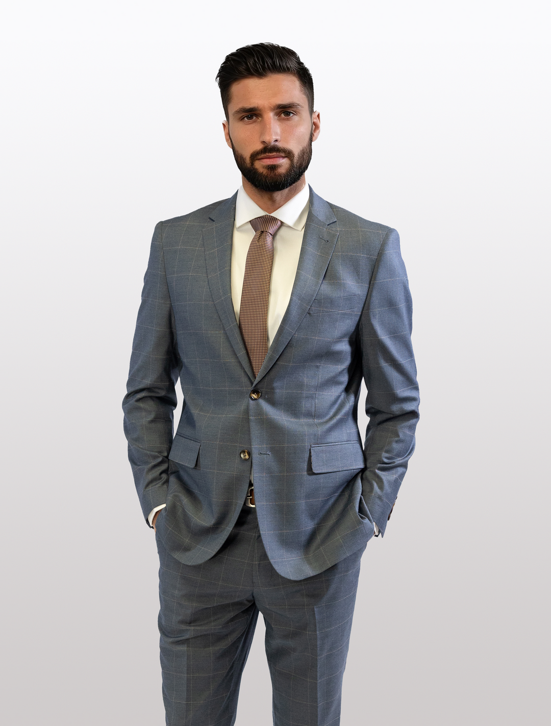 A man stands comfortably against a plain background, wearing the FUBU Gravel Blue 2-Piece Suit with a peach windowpane pattern, paired with a crisp white shirt and beige tie.