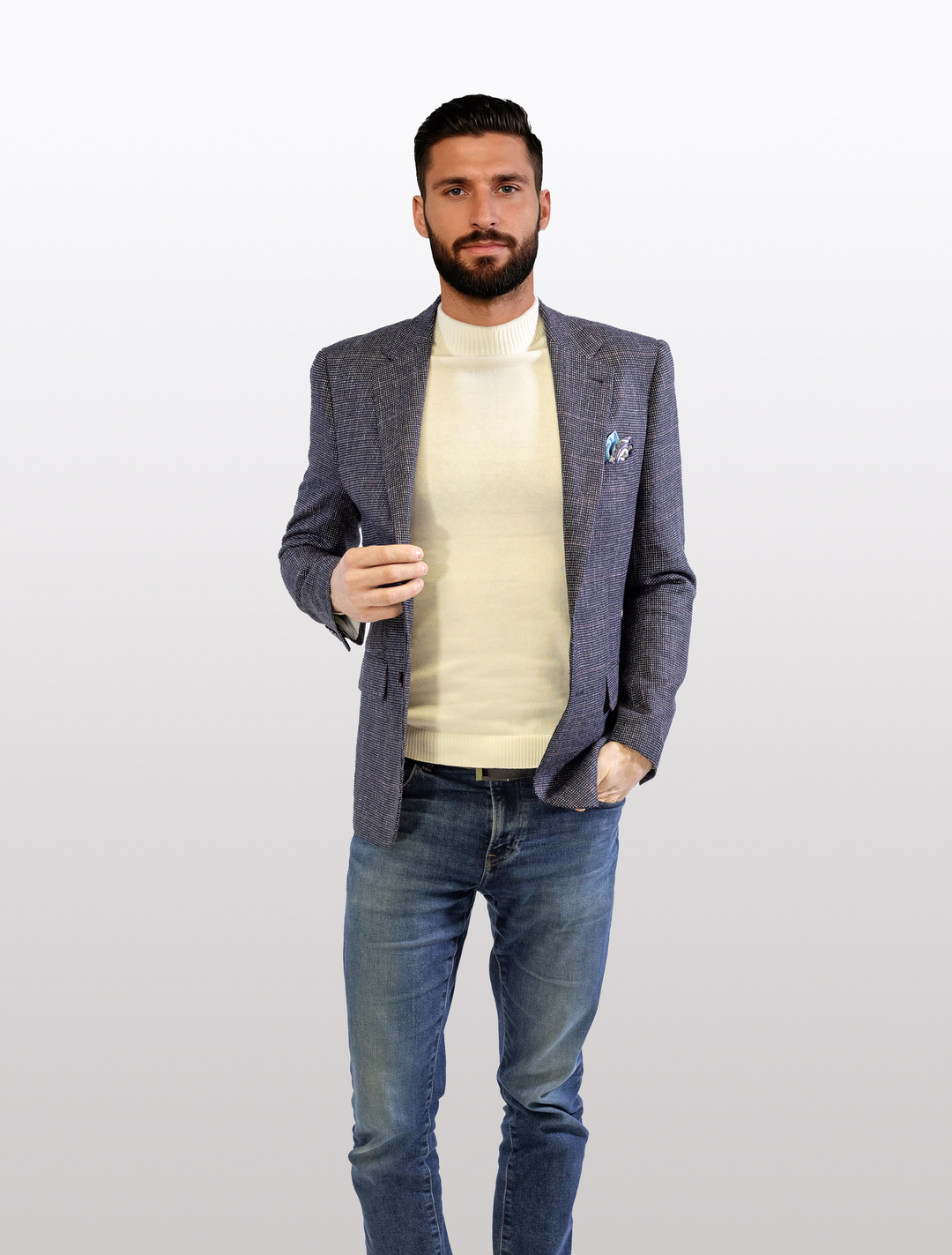 A man with a beard, dressed in a FUBU Textured Navy with Brick Windowpane Sport Jacket from Fubu The Colle, paired with a cream turtleneck and blue jeans, stands against a white background.