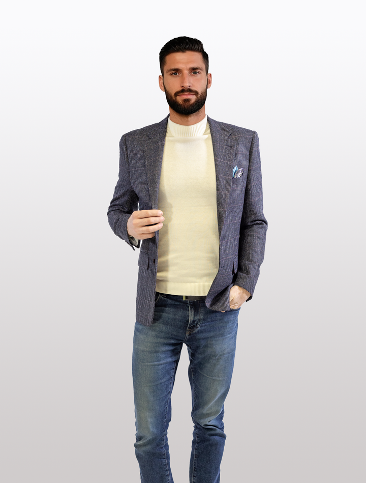 A man with a beard, dressed in a FUBU Textured Navy with Brick Windowpane Sport Jacket from Fubu The Colle, paired with a cream turtleneck and blue jeans, stands against a white background.
