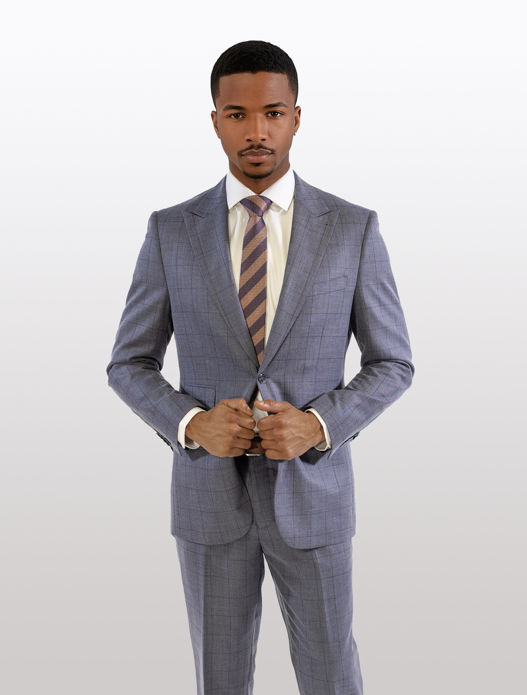 A man in a FUBU Wolf Grey with Rust Windowpane Peak Lapel 2-Piece Suit from Fubu The Colle stands against a plain background, buttoning his jacket over a crisp white shirt and striped tie.