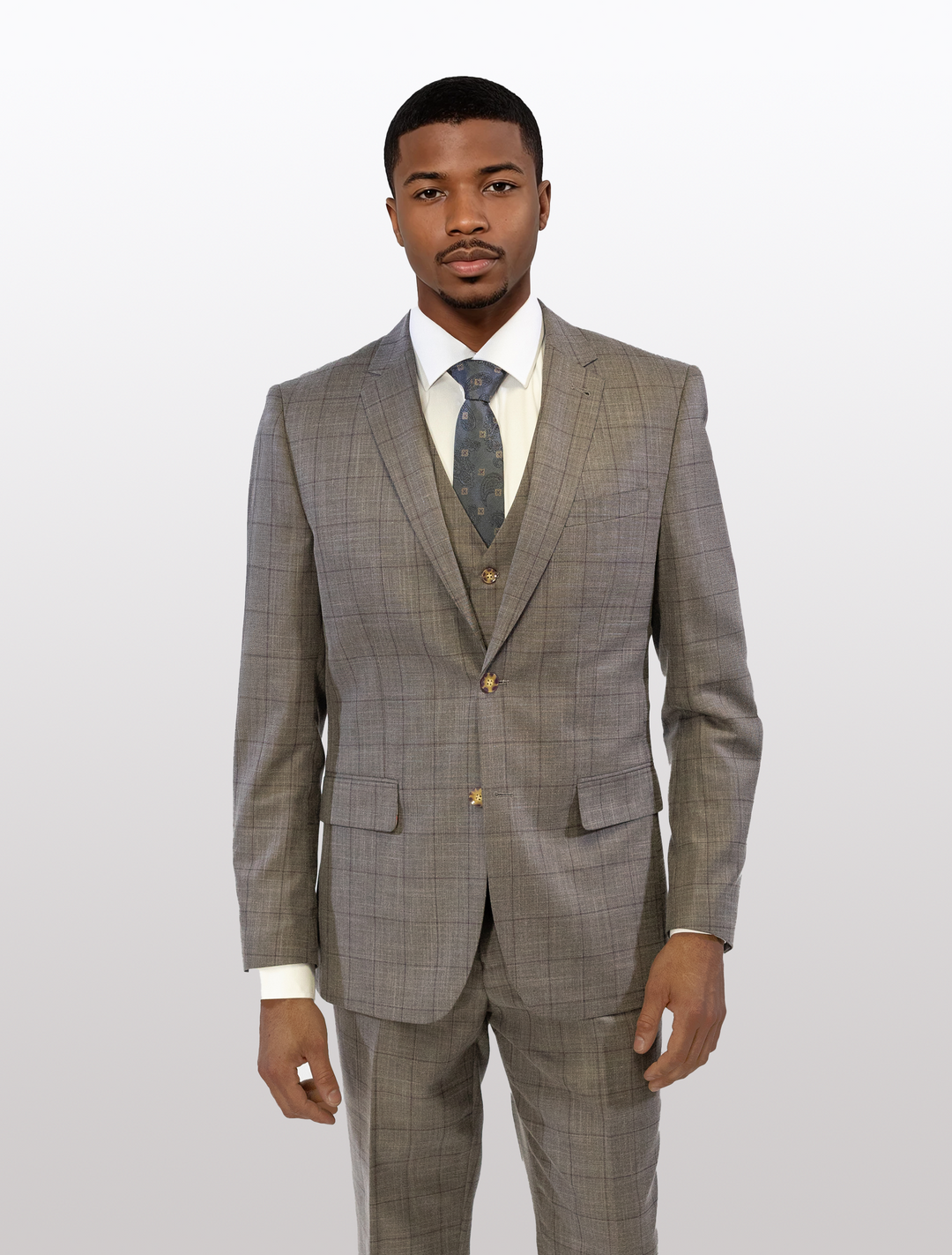 A man confidently stands against a plain background wearing a FUBU Taupe with Brown Windowpane Vested Suit by Fubu The Colle, complemented by a white shirt and patterned tie.