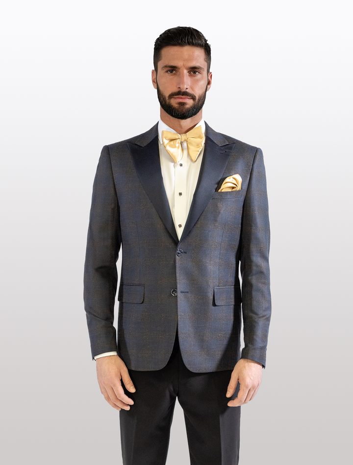 A bearded man stands against a plain background wearing a FUBU Navy with Bronze Windowpane Peak Lapel Tuxedo Jacket by Fubu The Colle, modern fit, complemented by a yellow bow tie and pocket square.