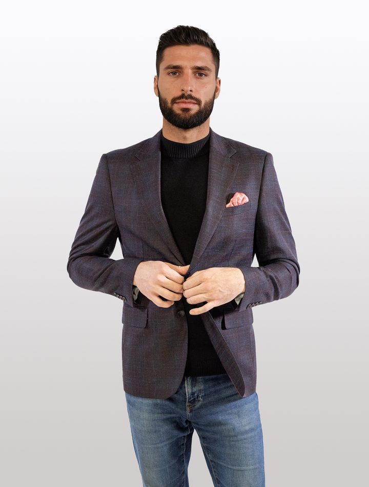 A bearded man, clad in a FUBU Midnight Navy with Wine Windowpane Sport Jacket from Fubu The Colle over a black sweater and jeans, stands against a light background. He adjusts his blazer, highlighting a bright pink pocket square with finesse.