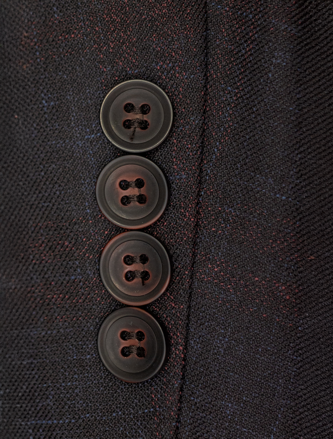 Close-up of four black buttons on a dark textured fabric, resembling those on the FUBU Midnight Navy with Wine Windowpane Sport Jacket by Fubu The Colle, arranged vertically along the edge.