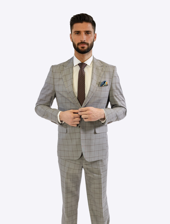 A man in a sleek FUBU Oat Grey with Brown Windowpane Peak Lapel 2-Piece Suit by Fubu The Colle, tailored with a slim fit, wears a white shirt and dark tie, buttoning his jacket against a plain background.