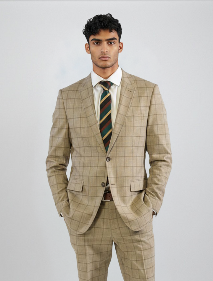 A man wearing the FUBU Camel with Mocha Windowpane 2-Piece Suit from Fubu The Colle, paired with a white shirt and striped tie, stands against a plain light gray background with his hands in his pockets.