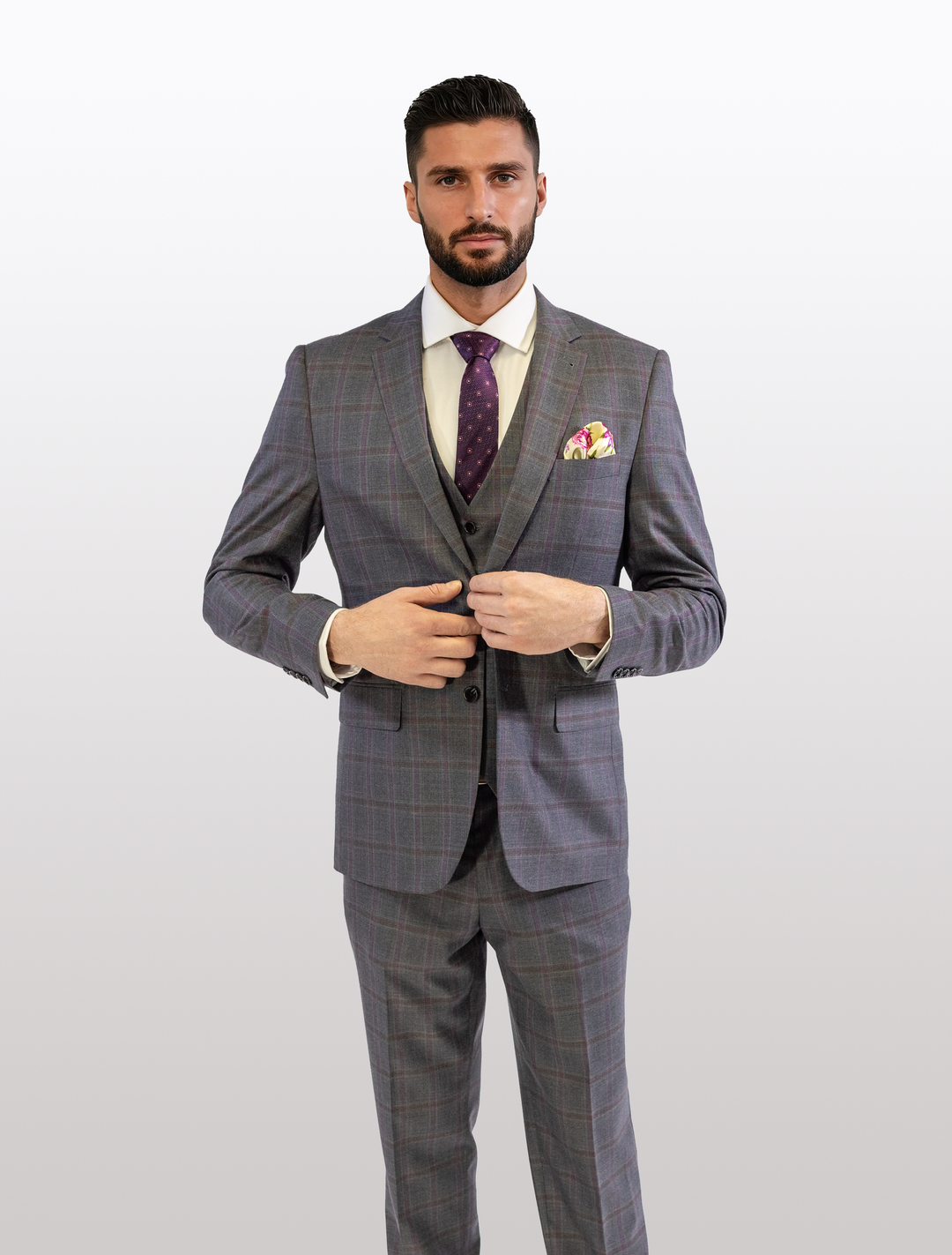 A man in a FUBU Charcoal with Purple Windowpane Vested Suit by Fubu The Colle, featuring a slim fit and stretch armhole, stands against a white background, exuding elegance and modernity with his matching purple tie and pocket square.