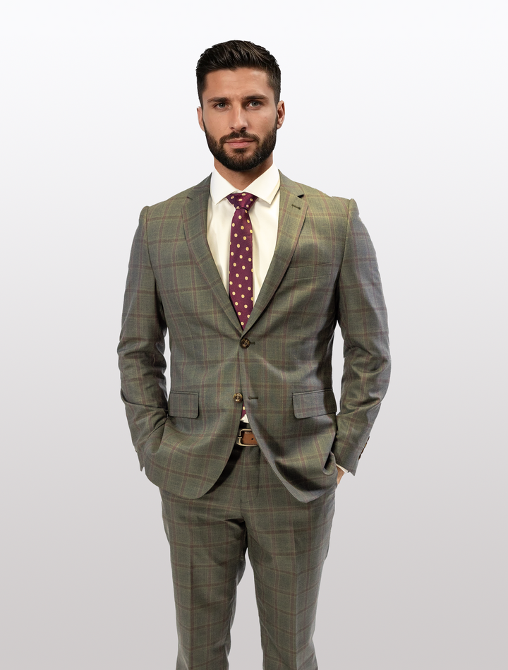 A dark-haired, bearded man stands with hands in pockets against a plain background wearing a FUBU Olive with Mahogany Windowpane 2-Piece Suit by Fubu The Colle, paired with a white shirt and polka dot tie.