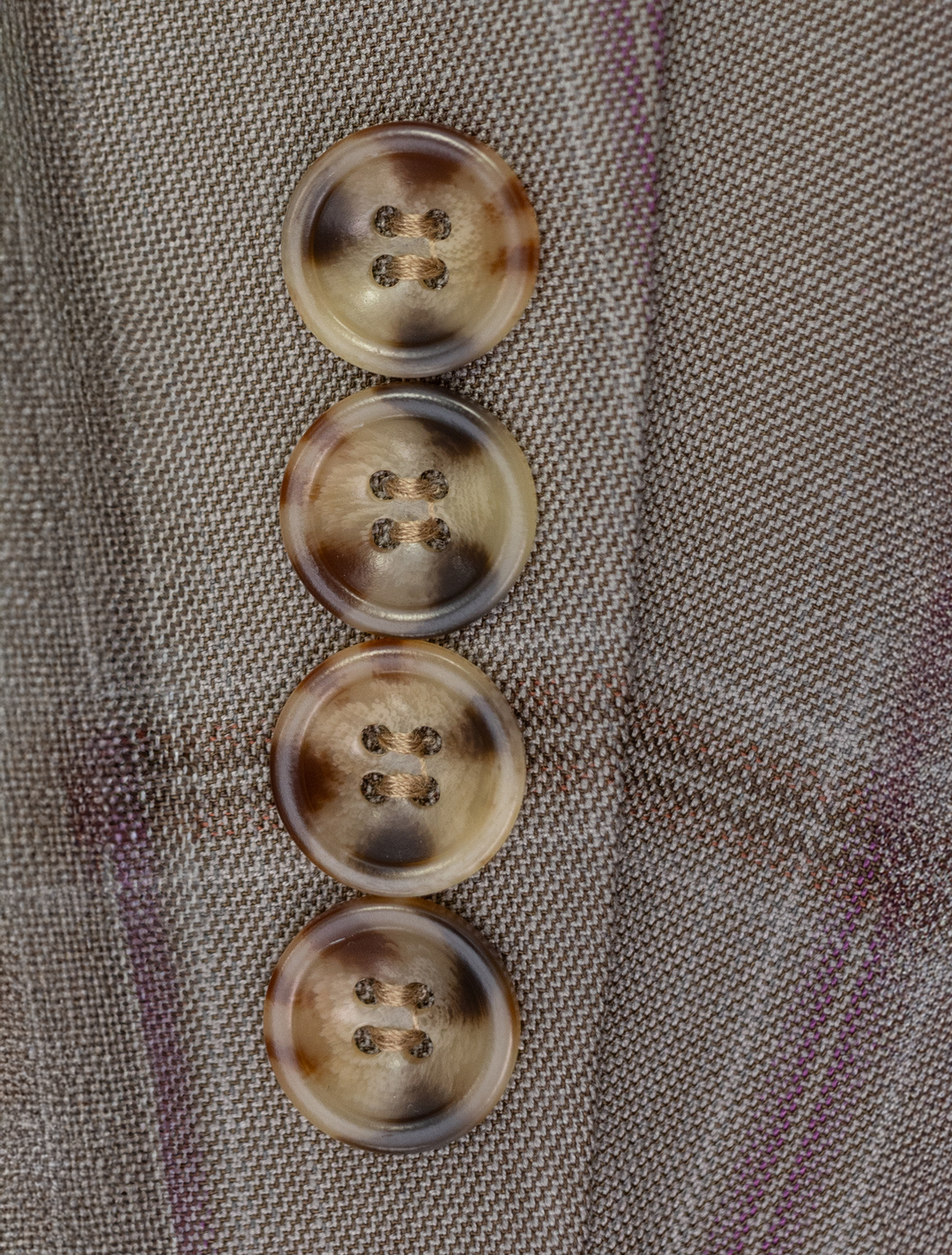 Close-up of four brown horn buttons on the FUBU Tan with Maroon Windowpane Vested Suit by Fubu The Colle, set vertically against a gray plaid fabric.