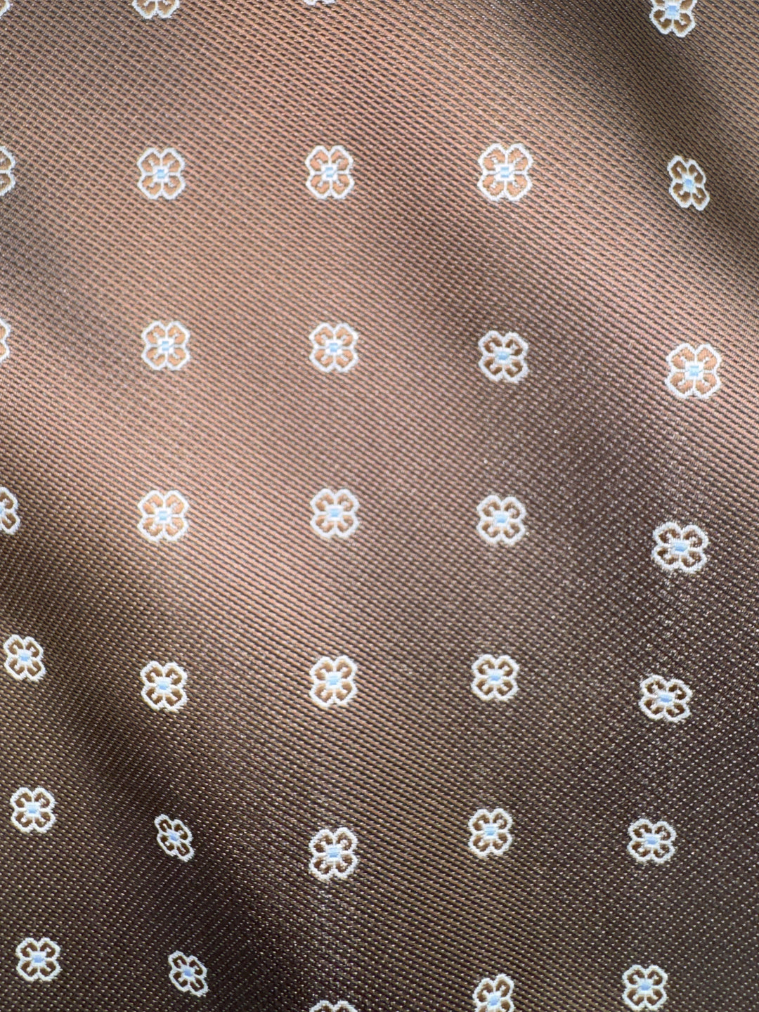 Close-up of the Karako Collecti Bronze Floral Pocket Square Handkerchief, showcasing a repeating grid pattern of small white outlined flowers on brown microfiber fabric.