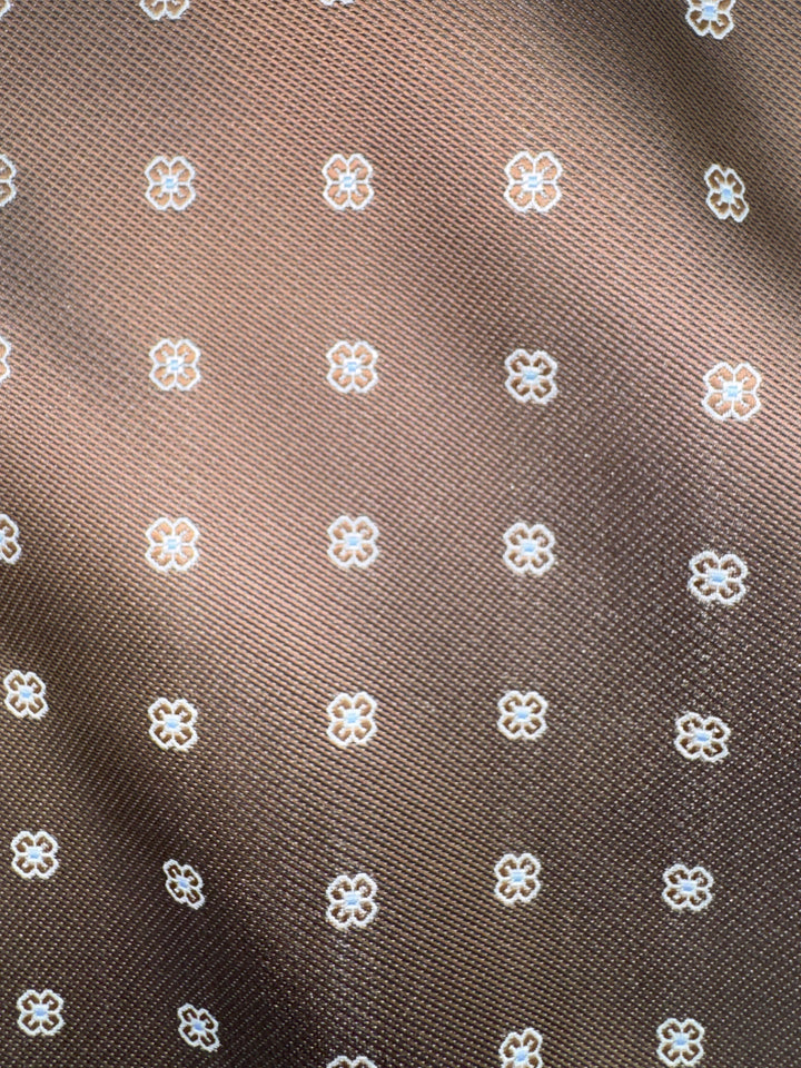Close-up of the Karako Collecti Bronze Floral Pocket Square Handkerchief, showcasing a repeating grid pattern of small white outlined flowers on brown microfiber fabric.