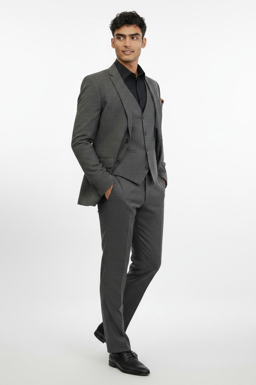 A man is wearing the FUBU Charcoal Grey Windowpane Vested Suit by Fubu The Colle, paired with a black shirt and black dress shoes. Standing with his hands in his pockets against a plain white background, he showcases this modern suit, which features a stretch armhole design for added comfort and ease of movement.