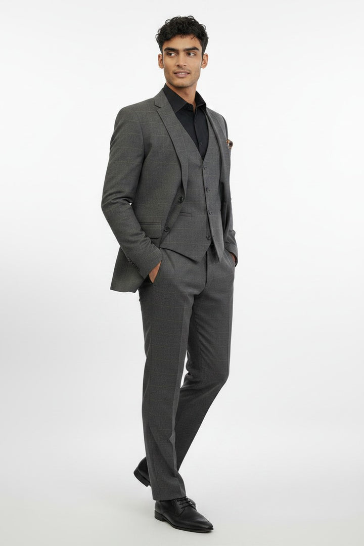 A man is wearing the FUBU Charcoal Grey Windowpane Vested Suit by Fubu The Colle, paired with a black shirt and black dress shoes. Standing with his hands in his pockets against a plain white background, he showcases this modern suit, which features a stretch armhole design for added comfort and ease of movement.