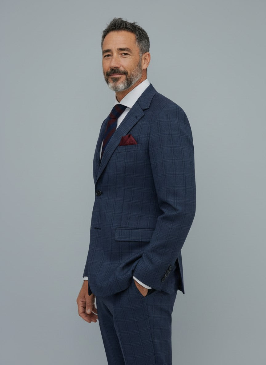 A man with gray hair and a beard wears the Gianco Ferro Men’s Dark Blue with Black Double Windowpane Slim Fit Suit, paired with a white shirt and dark tie, as he stands facing slightly to the side against a plain gray background.