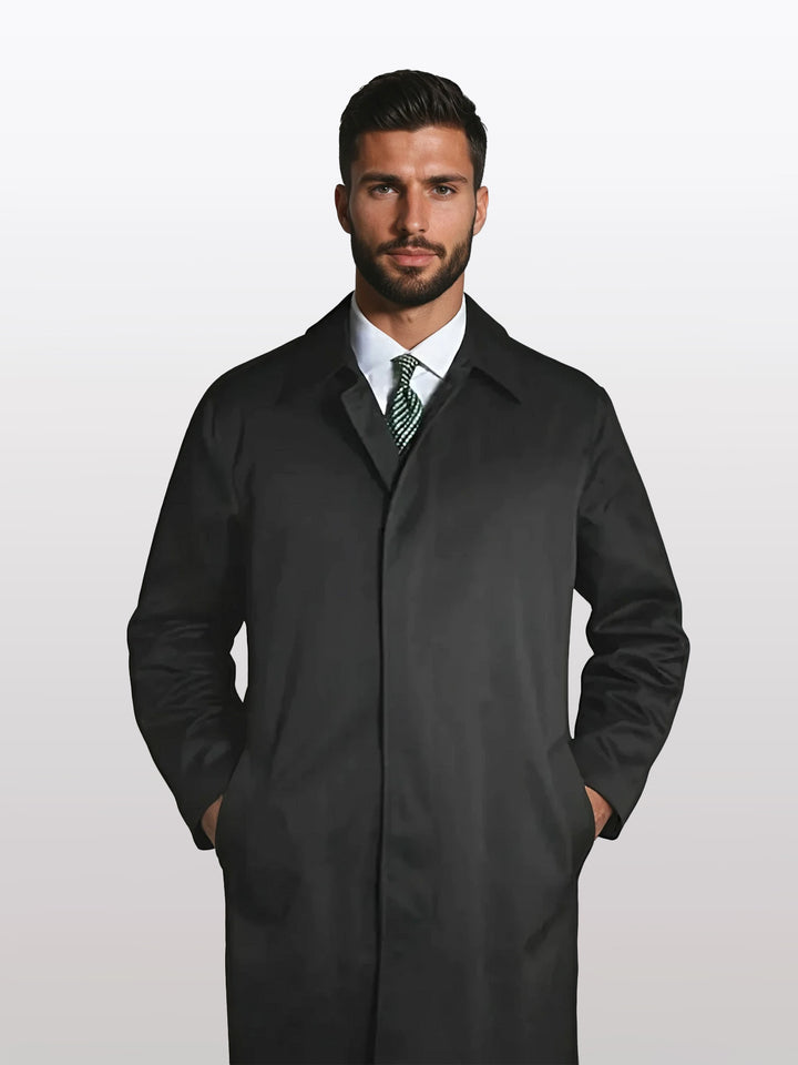 A man wearing a Jean Paul Germa Black Raincoat Jacket For Men - Full Length and Waterproof, with hands in pockets, white shirt, and green tie stands against a plain background.