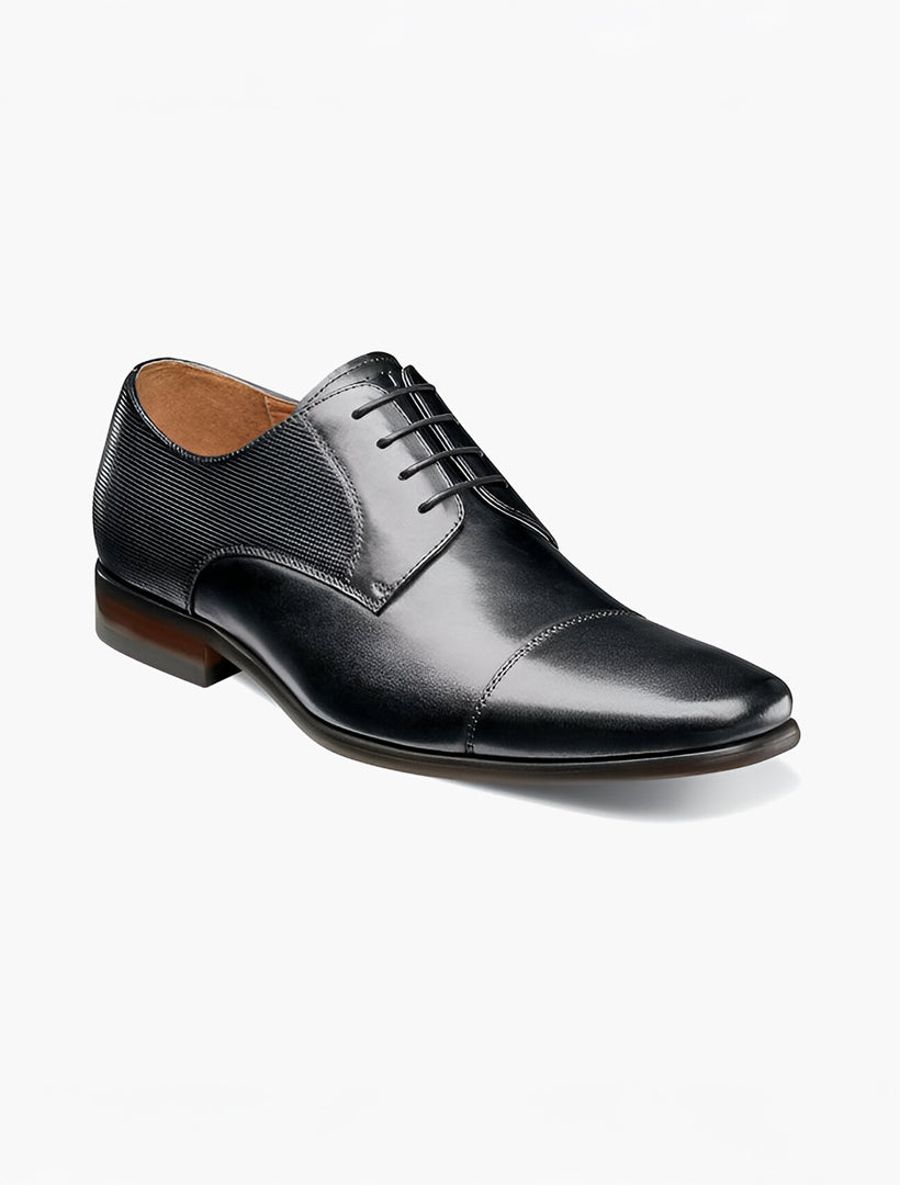 The Florsheim Black Cap Toe Oxford Shoe features a black leather upper, lace-up closure, and textured detail near the heel, combining classic design with contemporary elements. Displayed on a plain white background. Brand: Florsheim.