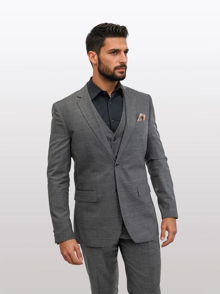 A man is wearing a FUBU Charcoal Grey Windowpane Vested Suit from Fubu The Colle, paired with a black shirt, against a plain background.