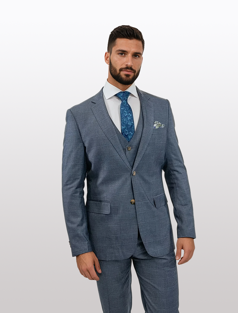 A man in a stylish FUBU Grey Windowpane Vested Suit by Fubu The Colle, accessorized with a patterned tie and pocket square, stands against a light gray background.