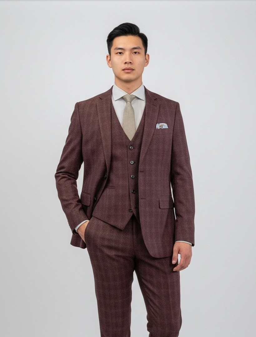 A man stands facing the camera wearing the FUBU Burgundy Tartan Vested Suit by Fubu The Colle, styled with a white shirt and gray tie against a plain light background. The modern-fit suit features a stretch armhole for added comfort and mobility.