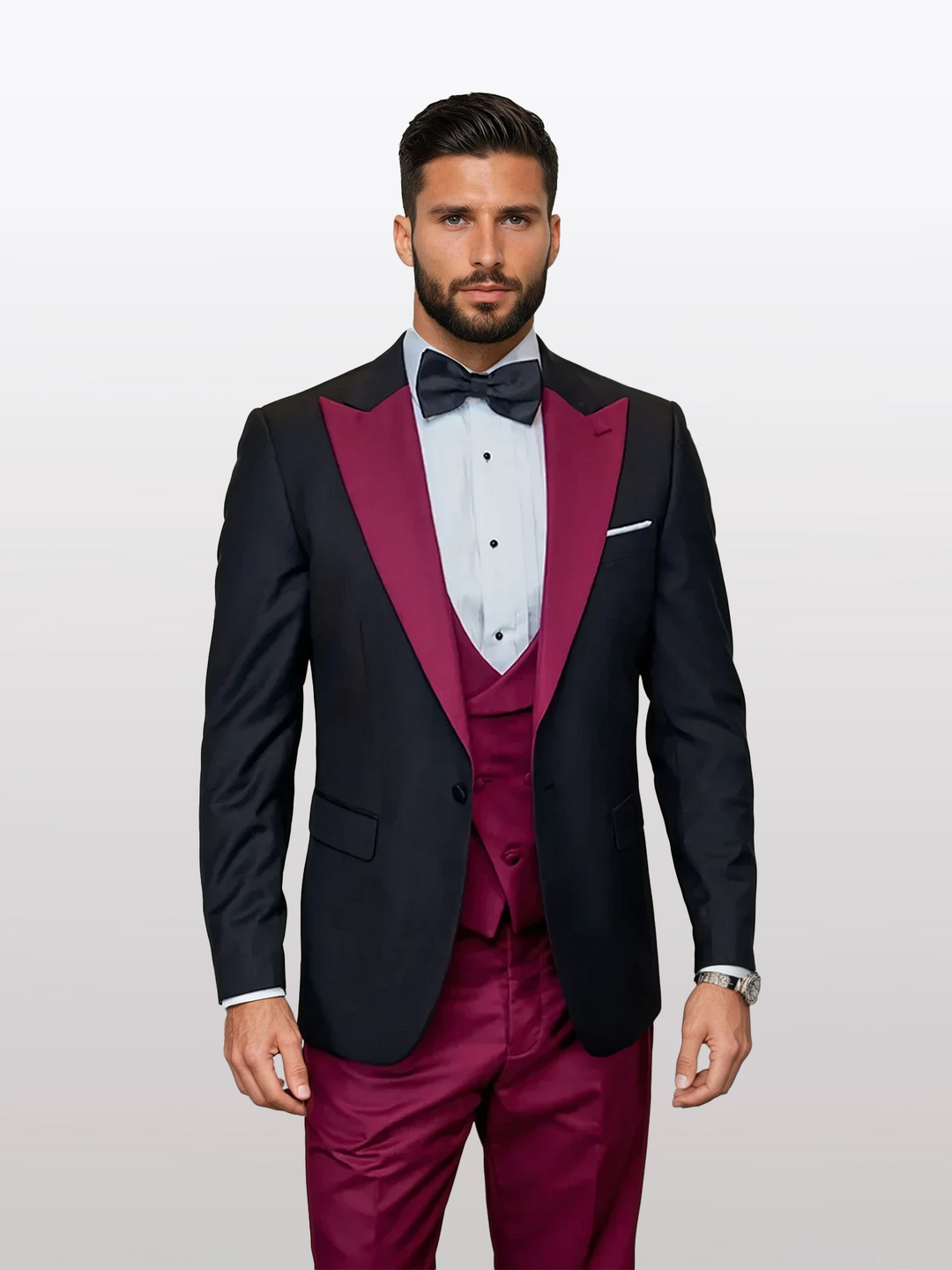 A man wearing the Statement Men's Black Vested Tuxedo with Burgundy Peak Lapel, made of 100% wool, complete with a bow tie and vest, stands against a light gray backdrop.