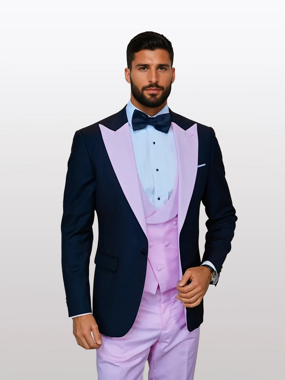 A man in the Statement Men's Black Vested Tuxedo, featuring a navy jacket with pastel pink peak lapels, a pink vest, and pink pants, stands against a light background. The ensemble is made from 100% wool for an elegant finish and complemented by a blue bow tie.