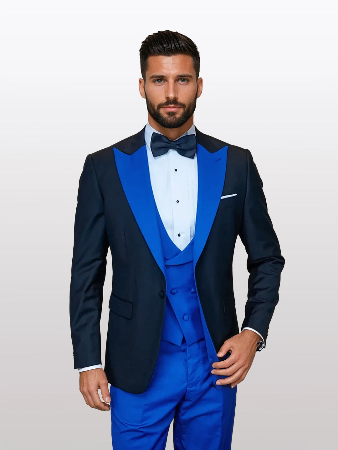 A man in a Statement Men's Black Vested Tuxedo with a Royal Blue Peak Lapel and 100% wool fabric, dons a black bow tie against a plain backdrop.