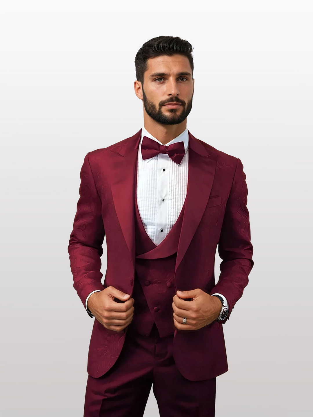 A man in a Statement Men's Burgundy Patterned Vested Tuxedo with Bowtie, made of 100% wool, adjusts his tailored fit jacket against a plain background.