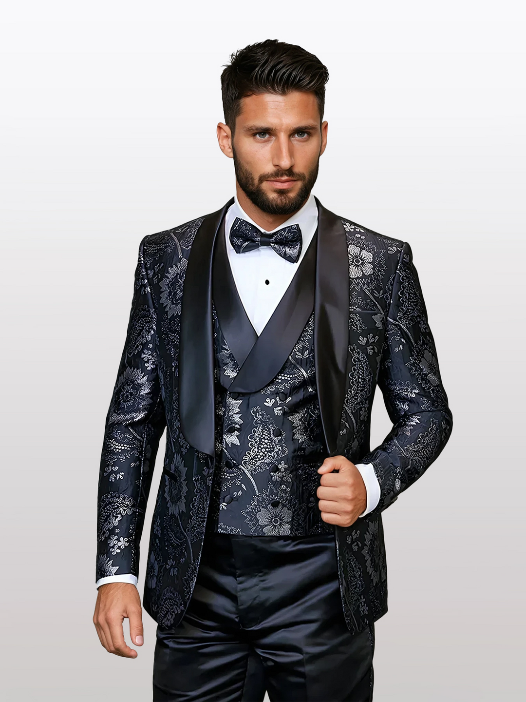 A man stands against a plain background in the Statement Men's Black Floral Patterned Vested Tuxedo, expertly crafted for a tailored fit. His ensemble features a black satin lapel, crisp white shirt, and bow tie.