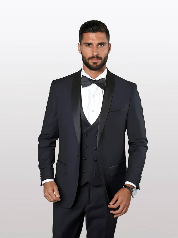 Man in a navy, Statement tailored-fit tuxedo with a shawl lapel and vest made of 100% wool, paired with a crisp white shirt and sleek black bow tie, posing against a plain background.