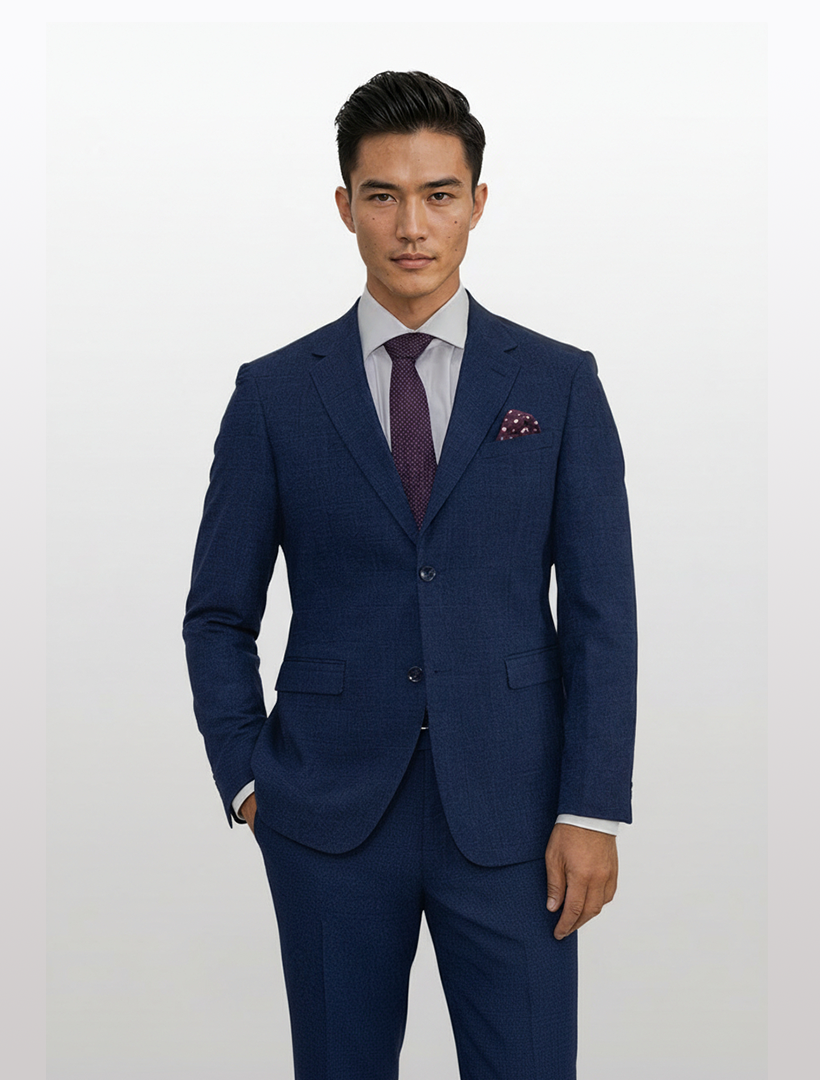 A man in a Gianco Ferro Men's Dark Blue With Red Windowpane Slim Fit Suit, paired with a white shirt and patterned purple tie, stands with one hand in his pocket against a plain light background.