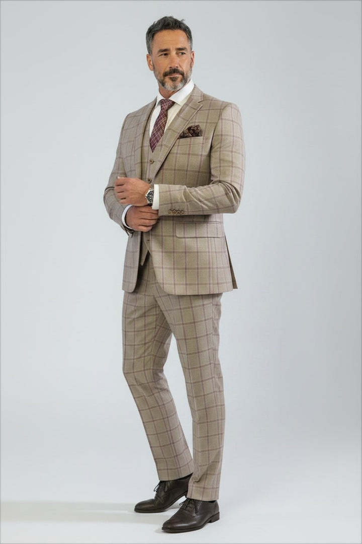 A man wearing the FUBU Tan with Maroon Windowpane Vested Suit by Fubu The Colle, paired with a white shirt, patterned tie, and dark brown shoes, stands against a plain light background.