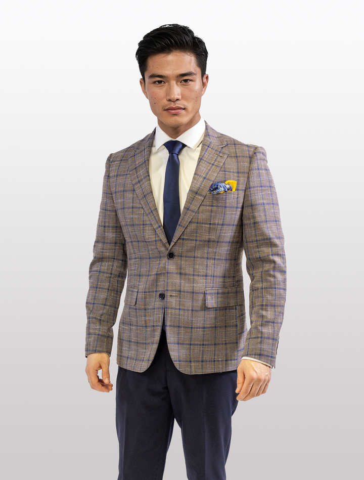 A man wearing a Fubu The Colle Cedar Brown Sport Jacket with Royal Blue Windowpane, paired with a white shirt and blue tie, stands against a plain background.