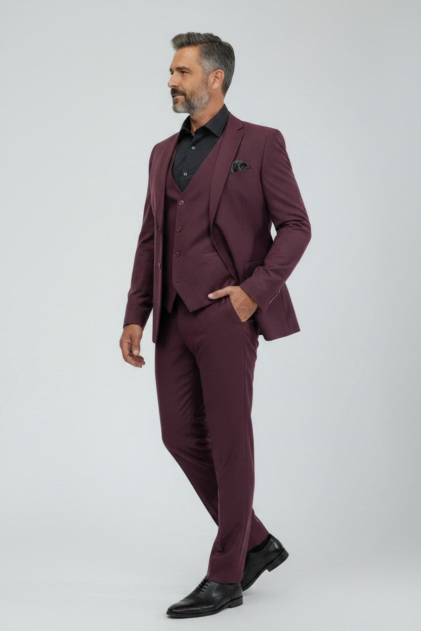 A man wearing the FUBU Burgundy Vested Suit by Fubu The Colle, paired with a black dress shirt and black shoes, stands against a plain light background looking to the left. The suit is designed with stretch armhole detailing for enhanced comfort and mobility.