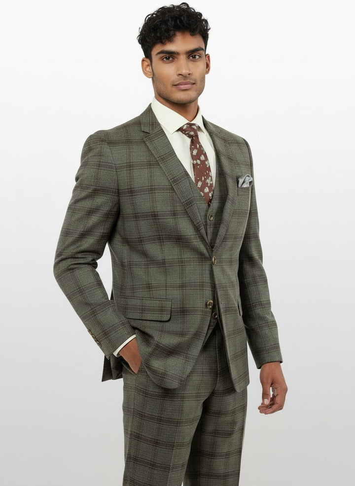 A man stands against a plain white background with one hand in his pocket, wearing the FUBU Charcoal with Brown Plaid Vested Suit by Fubu The Colle. The modern fit suit features stretch armholes for added comfort and ease of movement, paired with a cream shirt and a brown patterned tie.